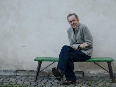 Kim Fupz Aakeson sidder udendørs på en grøn bænk foran en lys mur.